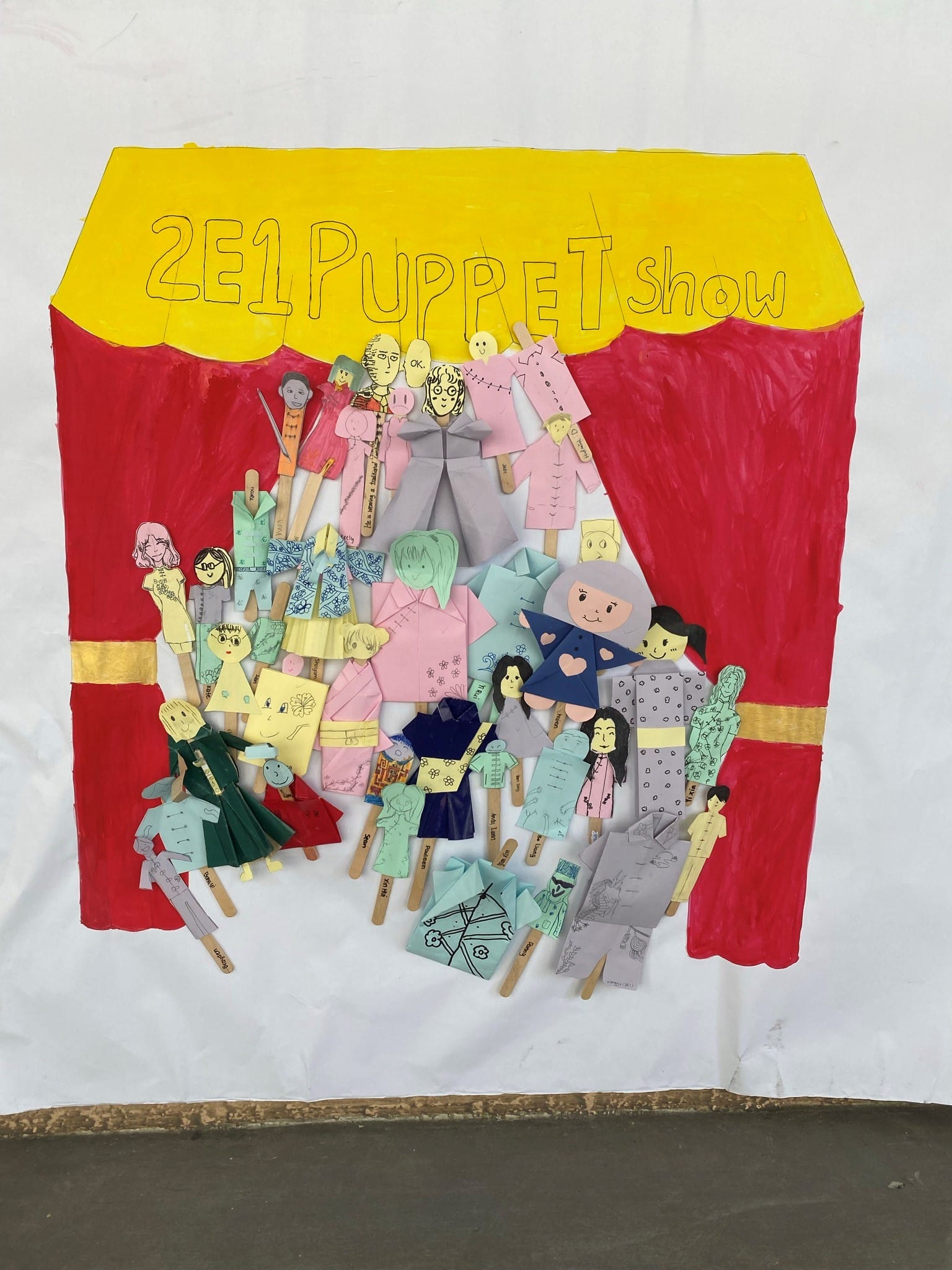 A hand-drawn stage with red curtains labeled "2E1 Puppet Show" displaying many colorful paper stick puppets.